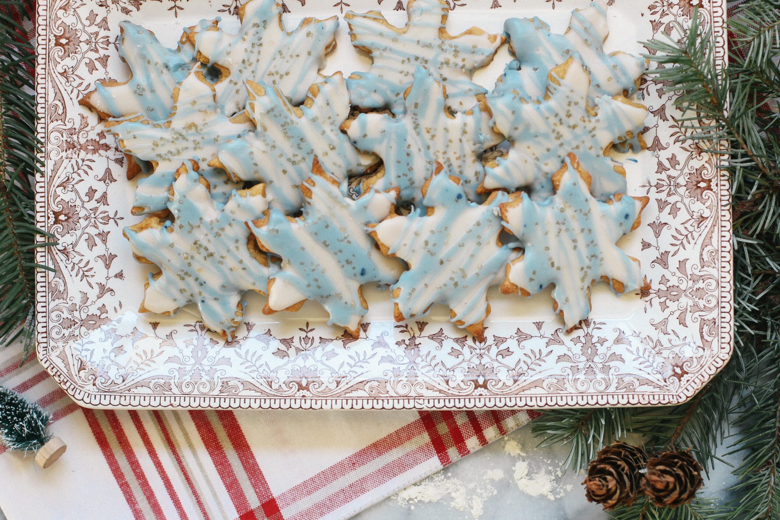 Photo of a tray of Einkorn Sugar Cookies in snowflake shapes with blue frosting and gold sprinkles on a transferware platter and christmas linens