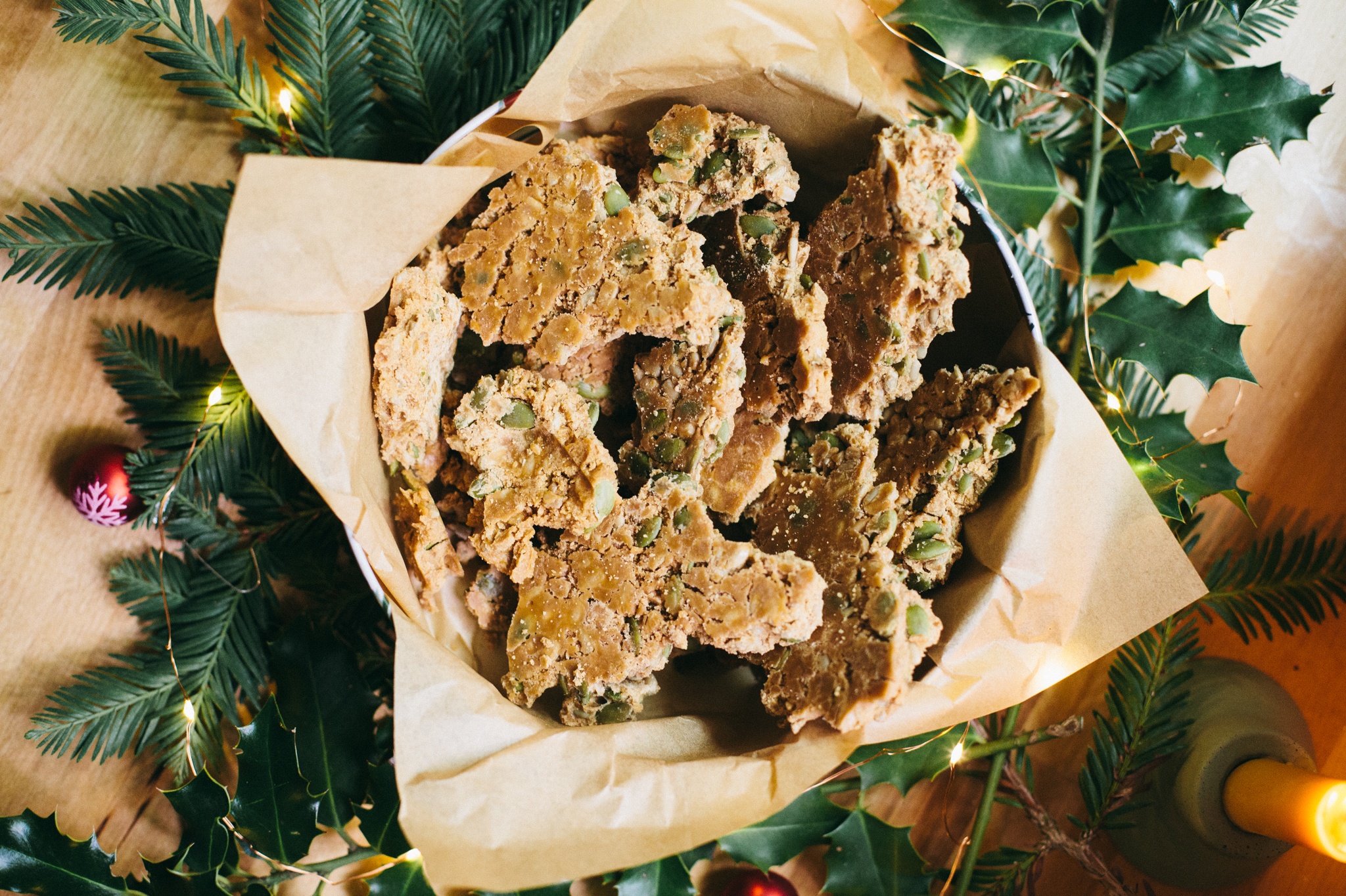 Photo of a tin of Maple Brittle in a holiday tin sitting in Christmas greens, lights and ornaments