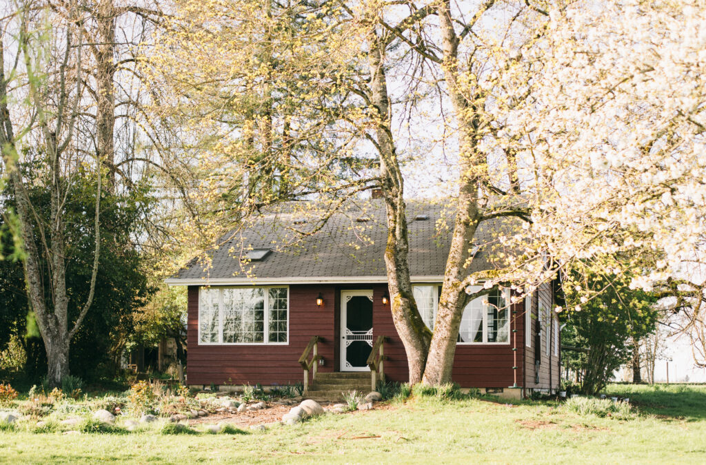 Photo of a red Swedish Cottage in Oregon surrounded by big trees and a cottage style garden with a rock path