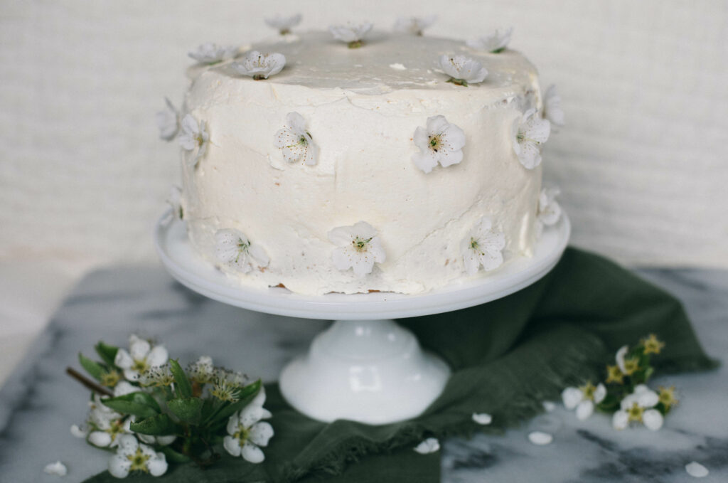 Photo of a white Einkorn Sponge Cake standing on a white cake stand covered in orchard blossoms