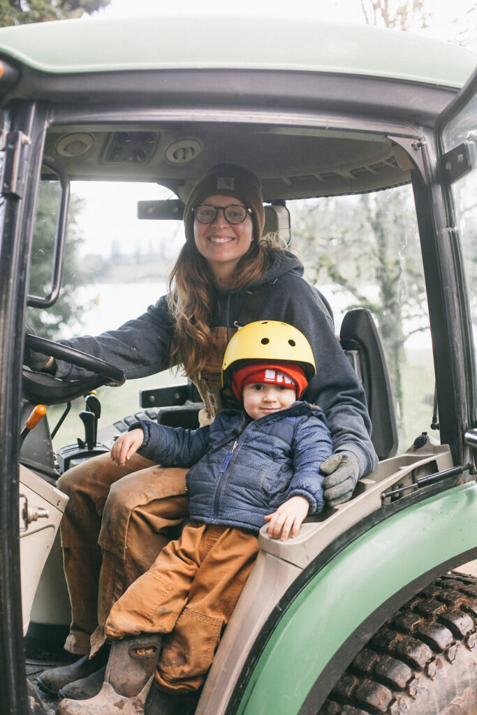 Image of a woman in Carhartt overalls in a John Deere tractor with her toddler boy working on a homestead