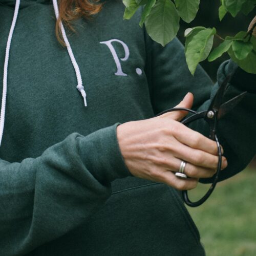 Image of a woman wearing a Potager Online green hoodie with an embroidered P trimming a fruit tree