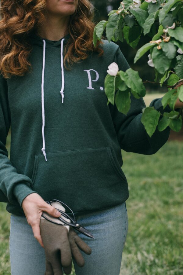 Image of a woman wearing a Potager Online green hoodie with an embroidered P trimming a fruit tree
