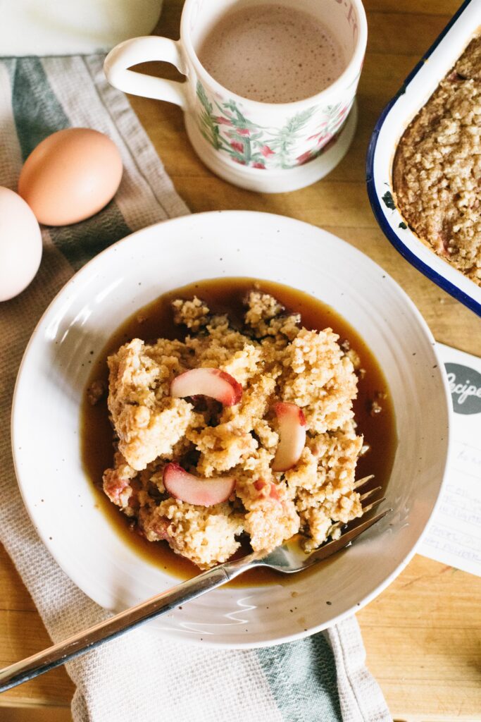 Photo of a bowl of Sourdough Rhubarb Baked Oatmeal in a white bowl with eggs, hot cocoa and a tea towel