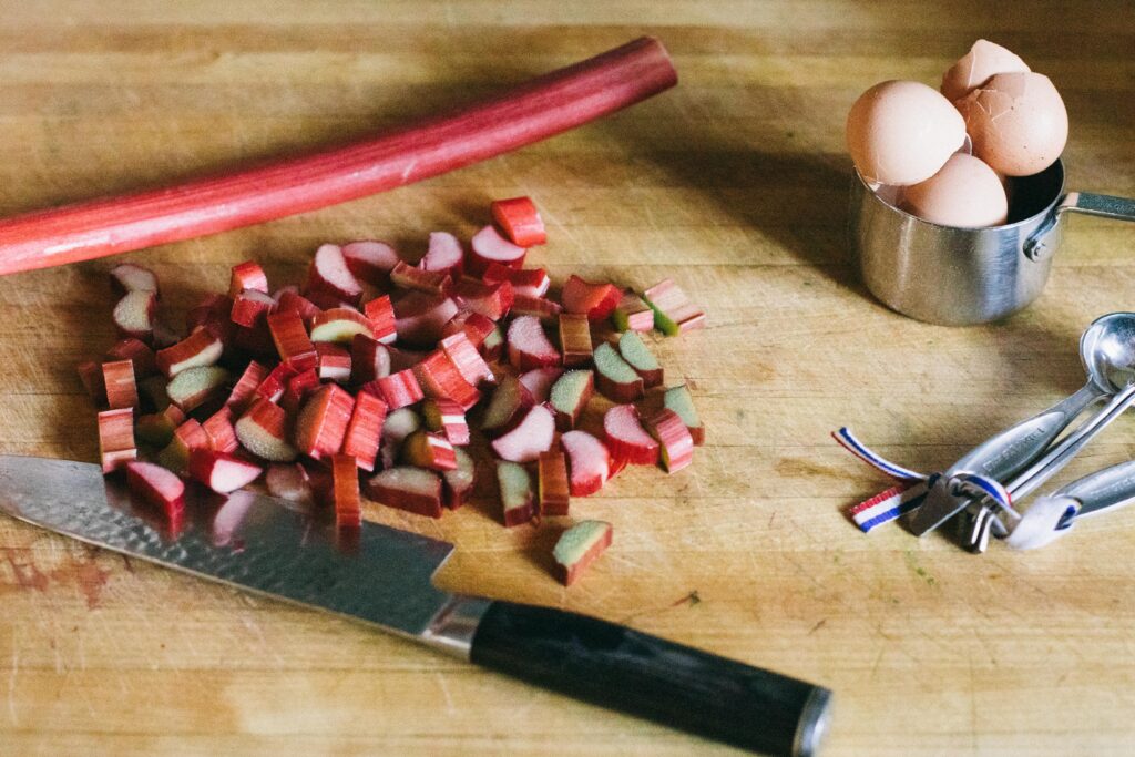 Photo of sliced Rhubarb sitting on a wooden cutting board with eggs and measuring spoons for a Sourdough Rhubarb Oatmeal Bake