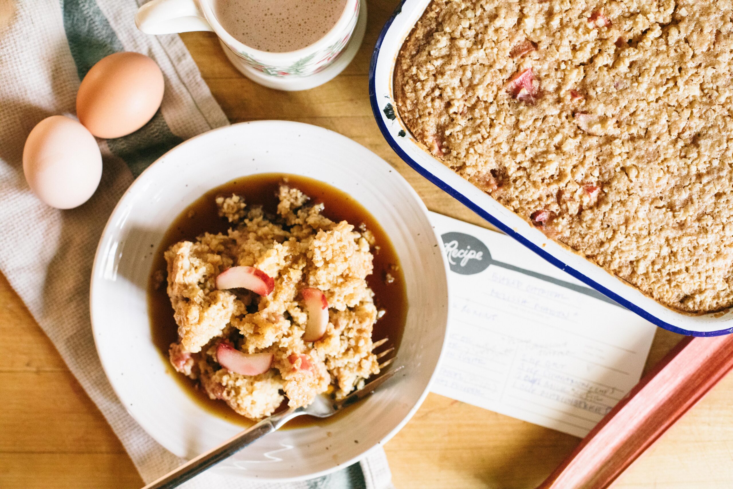 Photo of a bowl of Sourdough Rhubarb Baked Oatmeal with eggs, hot cocoa and rhubarb with a recipe card