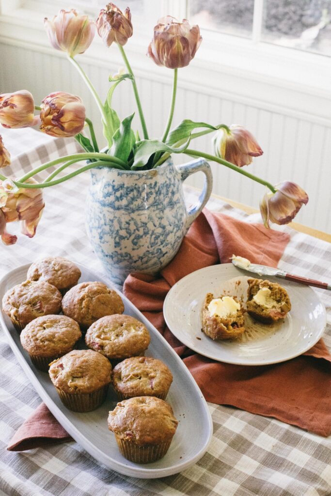 Image of a tray of rhubarb muffins on a gingham tablecloth with a spongeware pitcher of tulips on the dining room table in front of a white window