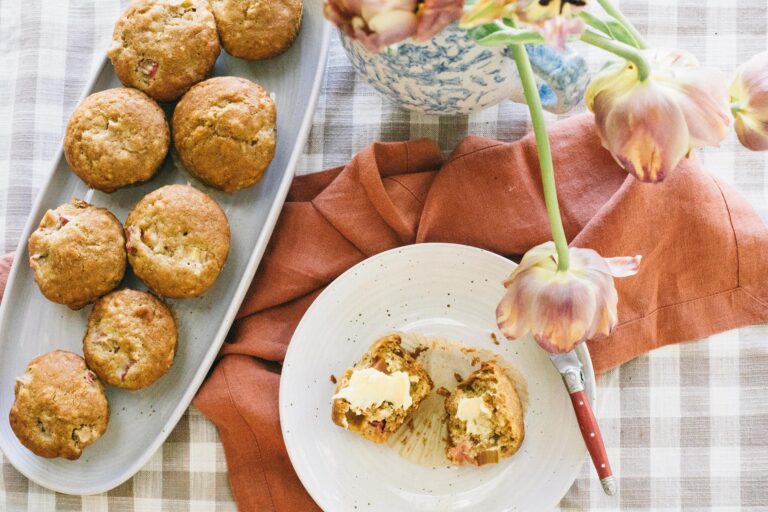 Photo of a a tray of rhubarb muffins on a gingham tablecloth with one muffin cut open and spread with butter