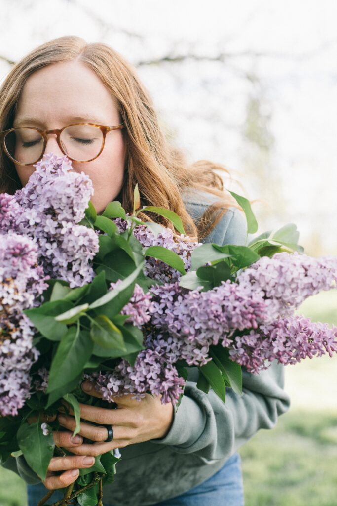 Image of a redheaded woman in glasses smelling a bunch of lilac flowers