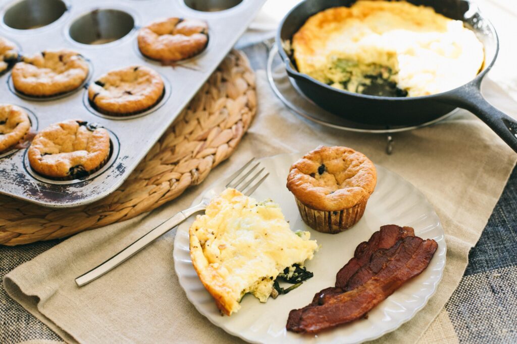 Image of an Einkorn Blueberry Muffin amidst a pan of muffins, frittata and bacon for breakfast